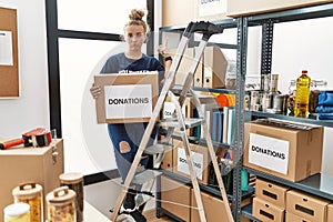 Young caucasian woman volunteer holding donations box pointing up looking sad and upset, indicating direction with fingers,