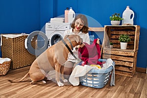 Young caucasian woman washing clothes sitting on floor with dog at laundry room