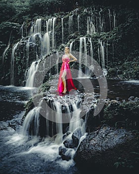 Young Caucasian woman at waterfall in tropical forest. Beautiful slim woman wearing long red dress. Banyu Wana Amertha waterfall