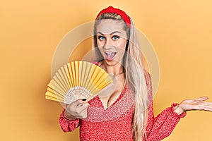 Young caucasian woman waving hand fan cooling air in summer celebrating achievement with happy smile and winner expression with