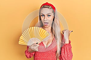 Young caucasian woman waving hand fan cooling air in summer pointing thumb up to the side smiling happy with open mouth