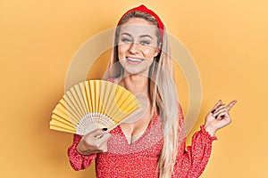 Young caucasian woman waving hand fan cooling air in summer smiling happy pointing with hand and finger to the side