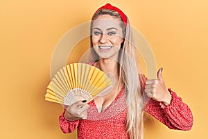 Young caucasian woman waving hand fan cooling air in summer smiling happy and positive, thumb up doing excellent and approval sign