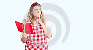 Young caucasian woman wearing apron holding recipe book and spoon serious face thinking about question with hand on chin,