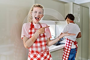 Young caucasian woman wearing apron and husband doing housework washing dishes amazed and smiling to the camera while presenting