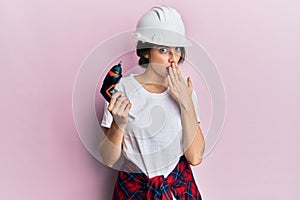 Young caucasian woman wearing architect hardhat using drill covering mouth with hand, shocked and afraid for mistake