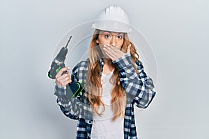 Young caucasian woman wearing architect hardhat using drill covering mouth with hand, shocked and afraid for mistake