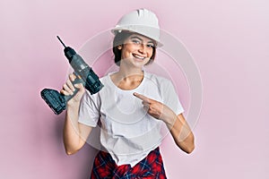 Young caucasian woman wearing architect hardhat using drill smiling happy pointing with hand and finger