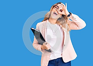 Young caucasian woman wearing business clothes and glasses holding binder smiling happy doing ok sign with hand on eye looking