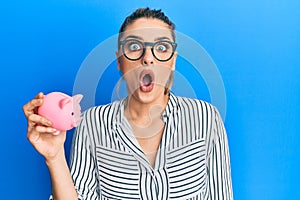 Young caucasian woman wearing business clothes holding piggy bank scared and amazed with open mouth for surprise, disbelief face