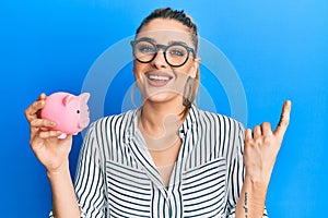 Young caucasian woman wearing business clothes holding piggy bank smiling happy pointing with hand and finger to the side