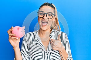Young caucasian woman wearing business clothes holding piggy bank smiling happy and positive, thumb up doing excellent and