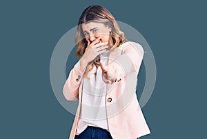 Young caucasian woman wearing business clothes laughing at you, pointing finger to the camera with hand over mouth, shame
