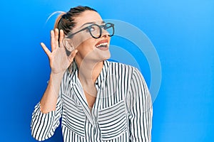 Young caucasian woman wearing business shirt and glasses smiling with hand over ear listening and hearing to rumor or gossip