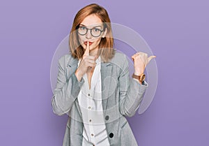 Young caucasian woman wearing business style and glasses asking to be quiet with finger on lips pointing with hand to the side