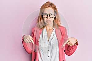 Young caucasian woman wearing business style and glasses pointing down looking sad and upset, indicating direction with fingers,