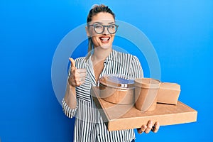 Young caucasian woman wearing business style holding delivery food smiling happy and positive, thumb up doing excellent and