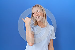 Young caucasian woman wearing casual blue t shirt smiling with happy face looking and pointing to the side with thumb up