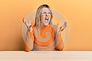 Young caucasian woman wearing casual clothes and glasses sitting on the table crazy and mad shouting and yelling with aggressive