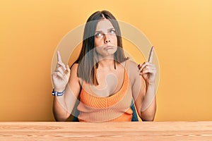 Young caucasian woman wearing casual clothes sitting on the table pointing up looking sad and upset, indicating direction with