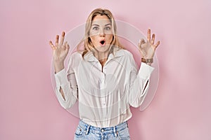 Young caucasian woman wearing casual white shirt over pink background looking surprised and shocked doing ok approval symbol with