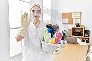 Young caucasian woman wearing cleaner uniform holding cleaning products cleaning office with open hand doing stop sign with