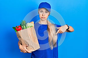 Young caucasian woman wearing courier uniform with groceries from supermarket pointing down looking sad and upset, indicating