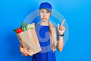 Young caucasian woman wearing courier uniform with groceries from supermarket pointing up looking sad and upset, indicating