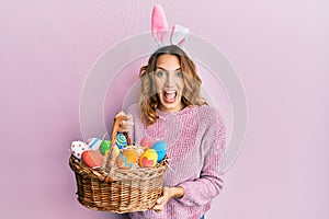 Young caucasian woman wearing cute easter bunny ears holding colored egg celebrating crazy and amazed for success with open eyes