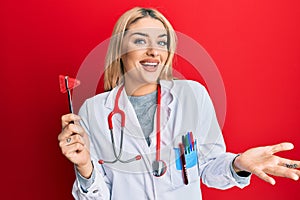 Young caucasian woman wearing doctor uniform holding medical reflex hammer celebrating achievement with happy smile and winner