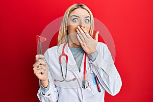 Young caucasian woman wearing doctor uniform holding medical reflex hammer covering mouth with hand, shocked and afraid for