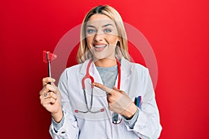 Young caucasian woman wearing doctor uniform holding medical reflex hammer smiling happy pointing with hand and finger