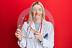 Young caucasian woman wearing doctor uniform holding tuning fork sticking tongue out happy with funny expression