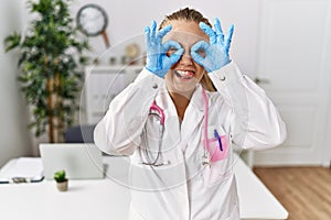 Young caucasian woman wearing doctor uniform and stethoscope at the clinic doing ok gesture like binoculars sticking tongue out,