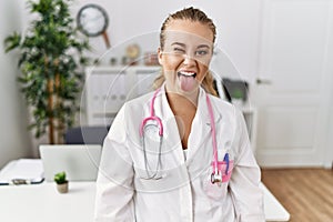 Young caucasian woman wearing doctor uniform and stethoscope at the clinic sticking tongue out happy with funny expression