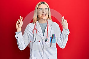 Young caucasian woman wearing doctor uniform and stethoscope gesturing finger crossed smiling with hope and eyes closed