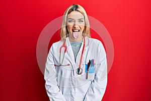 Young caucasian woman wearing doctor uniform and stethoscope sticking tongue out happy with funny expression