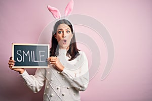 Young caucasian woman wearing easter rabbit ears and holding blackboard with smile word scared in shock with a surprise face,
