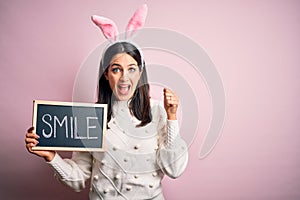 Young caucasian woman wearing easter rabbit ears and holding blackboard with smile word screaming proud and celebrating victory
