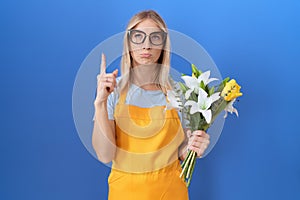 Young caucasian woman wearing florist apron holding flowers pointing up looking sad and upset, indicating direction with fingers,