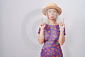 Young caucasian woman wearing flowers dress and summer hat pointing up looking sad and upset, indicating direction with fingers,