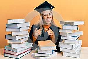 Young caucasian woman wearing graduation ceremony robe sitting on the table pointing fingers to camera with happy and funny face