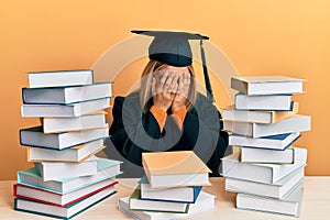 Young caucasian woman wearing graduation ceremony robe sitting on the table with sad expression covering face with hands while