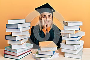 Young caucasian woman wearing graduation ceremony robe sitting on the table tired hands covering face, depression and sadness,