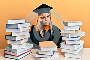 Young caucasian woman wearing graduation ceremony robe sitting on the table worried and stressed about a problem with hand on