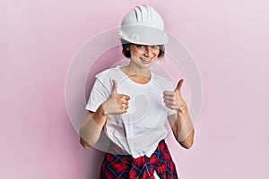 Young caucasian woman wearing hardhat success sign doing positive gesture with hand, thumbs up smiling and happy