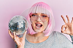 Young caucasian woman wearing pink wig holding disco ball doing ok sign with fingers, smiling friendly gesturing excellent symbol