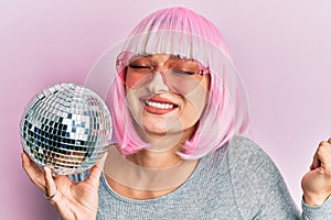 Young caucasian woman wearing pink wig holding disco ball screaming proud, celebrating victory and success very excited with