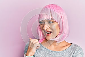 Young caucasian woman wearing pink wig smiling with happy face looking and pointing to the side with thumb up