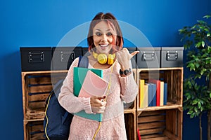 Young caucasian woman wearing student backpack and books pointing thumb up to the side smiling happy with open mouth